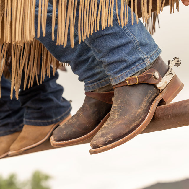 Close up photo of three people sitting on a gate wearing cowboy boots.